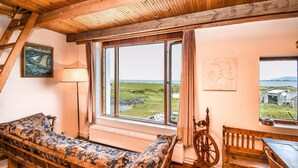 Ein rustikaler Raum mit Holzbalkendecke, einem großen Fenster mit Blick auf die Landschaft, einer hölzernen Bank und einem Spinnrad.