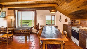 Ein rustikaler Essbereich mit Holztisch und Bänken, große Fenster mit Blick auf die Landschaft, und eine Küche mit Holzschränken und einem Herd.