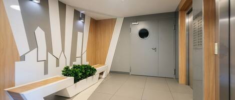 Un área de recepción moderna con un banco, una planta en maceta, una lámpara de pared y una puerta con un ventanal redondo.