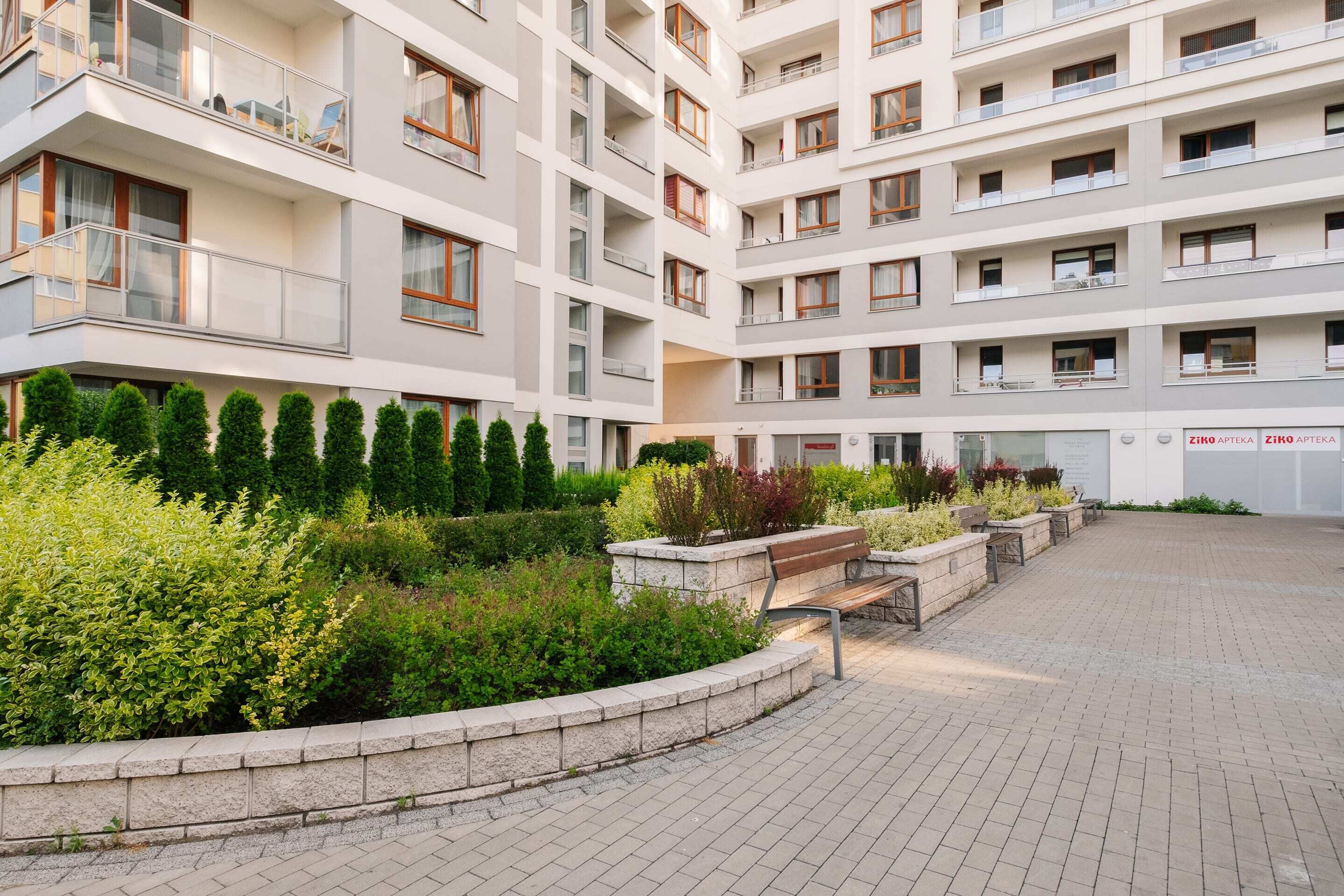 A multi-story residential building with balconies, a landscaped garden area with a bench, and a paved walkway.