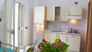 A compact kitchen with white cabinets, a sink, and a stove. There is a dining table with a checkered tablecloth and a vase with flowers. - Villaggio Turistico Defensola (Vieste)