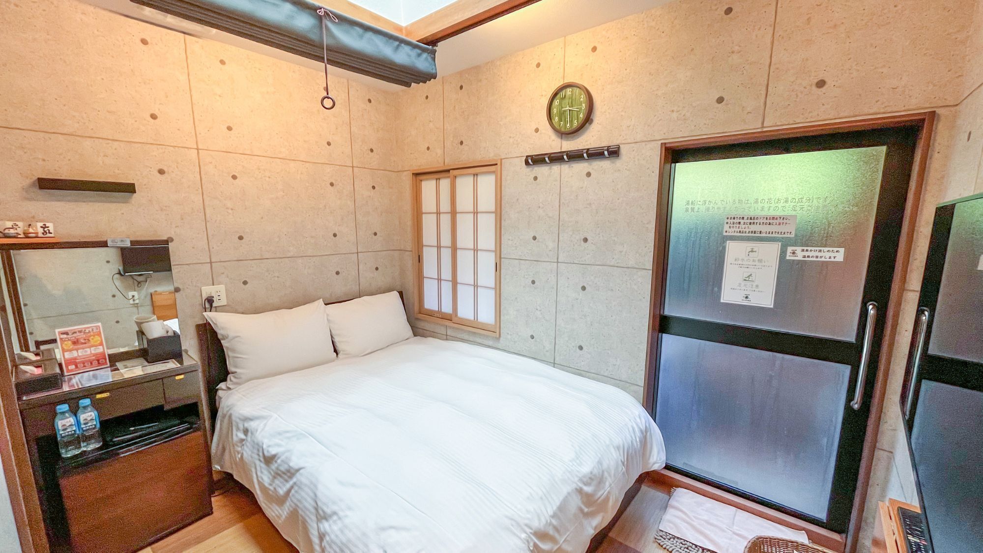 A hotel room with a bed, a mirror, a clock, a window, and a refrigerator.