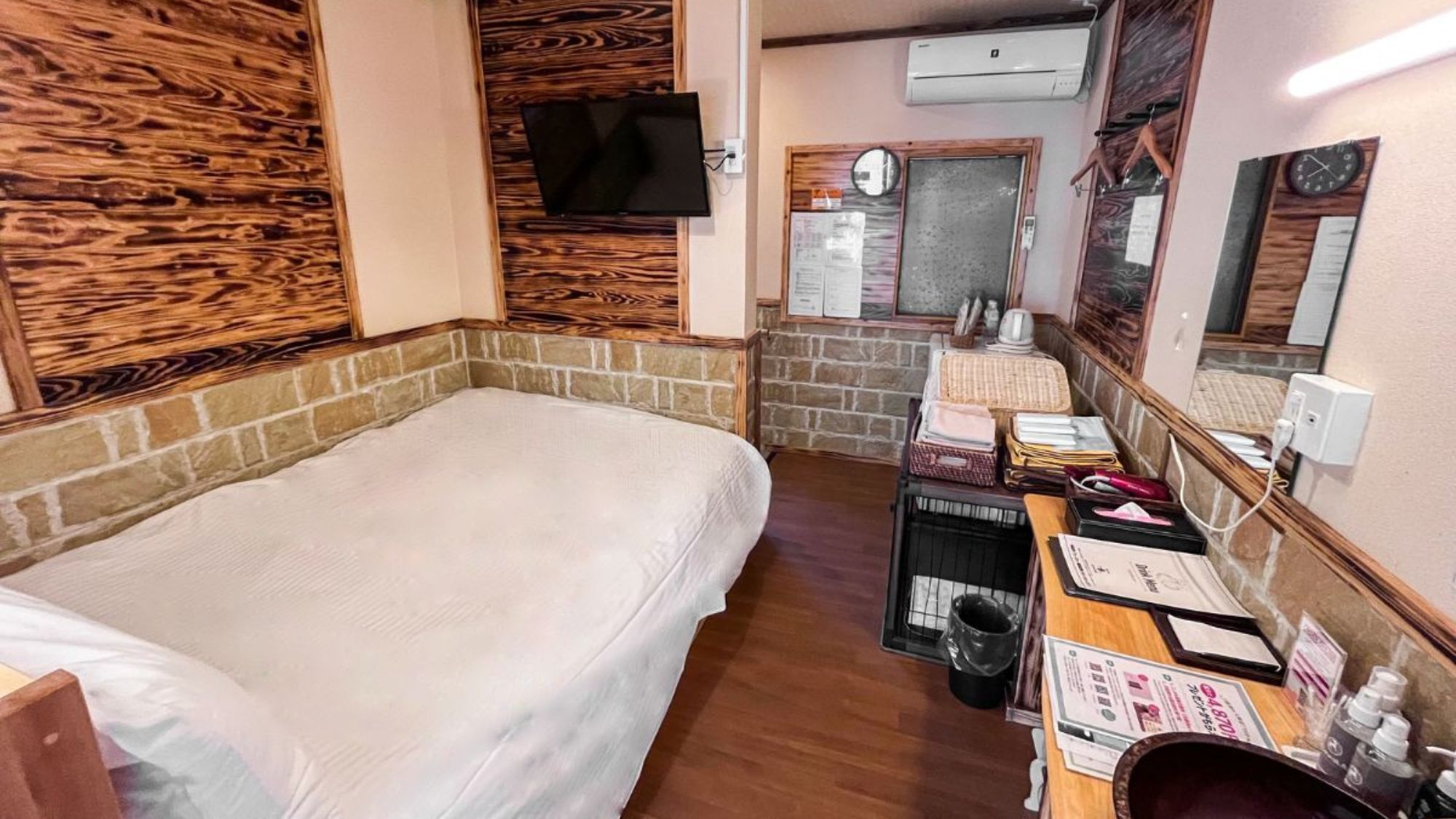 A room with a bed, a desk with papers, a trash bin, and a wall-mounted TV.