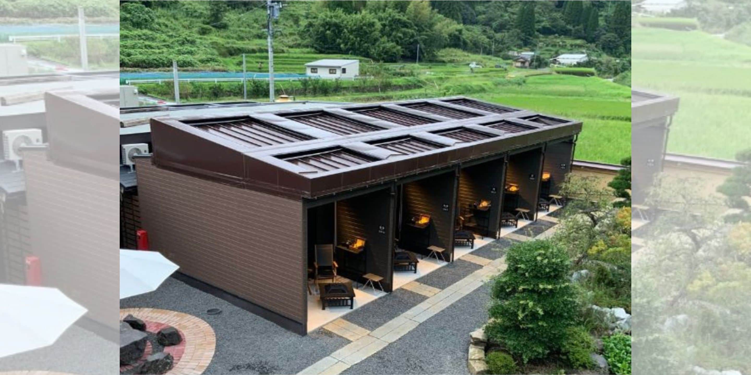 A modern building with a flat roof, outdoor seating area with tables and chairs, and a view of a green landscape with fields and trees.