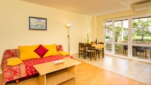 A living room with a sofa, coffee table, dining table, chairs, and a large window with a view of greenery. - Apartments Radic (Dubrovnik)