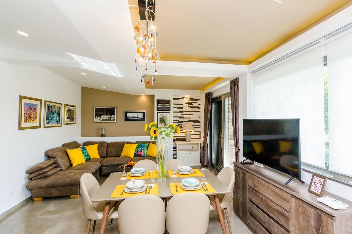 Un salon moderne comprenant un coin repas, un ensemble de canapés, un téléviseur et un lustre décoratif.