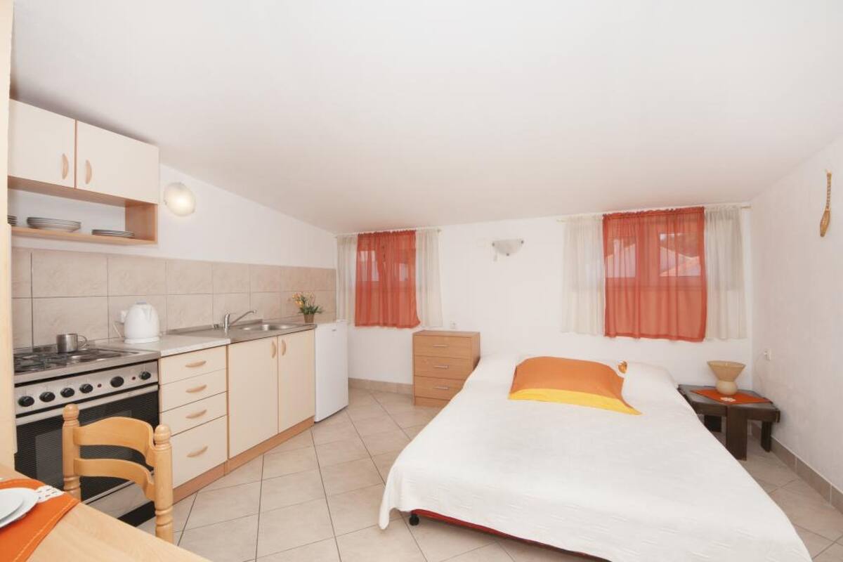 A compact room with a kitchenette on one side, a bed with an orange accent, and a small bedside table.