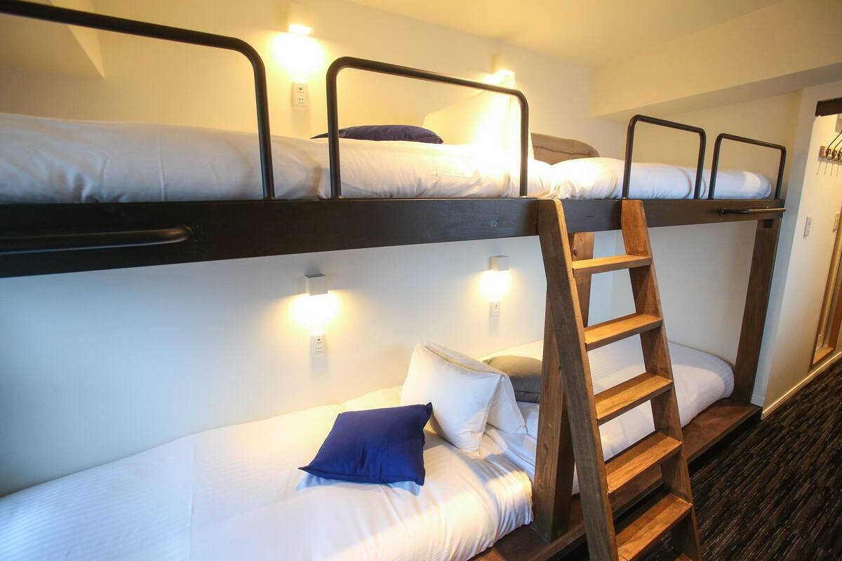 Une chambre avec un lit superposé, une échelle en bois, du linge de lit blanc et des lampes fixées au mur.