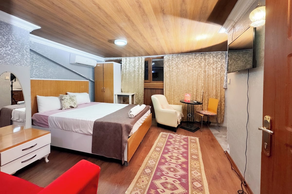 A hotel room with a bed, a desk, a chair, a small table, and a TV mounted on the wall.