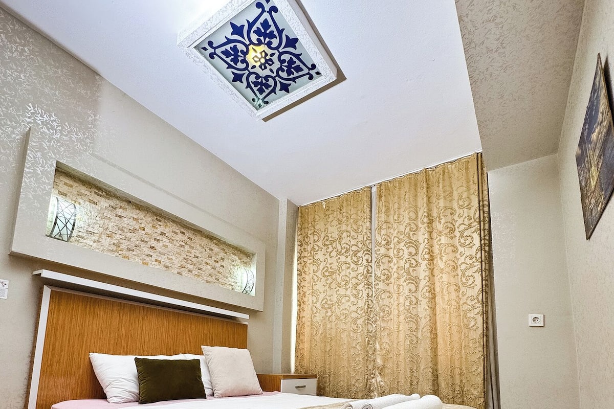 A bedroom with a bed, a nightstand, a window with curtains, a wall-mounted light fixture, and a ceiling light.