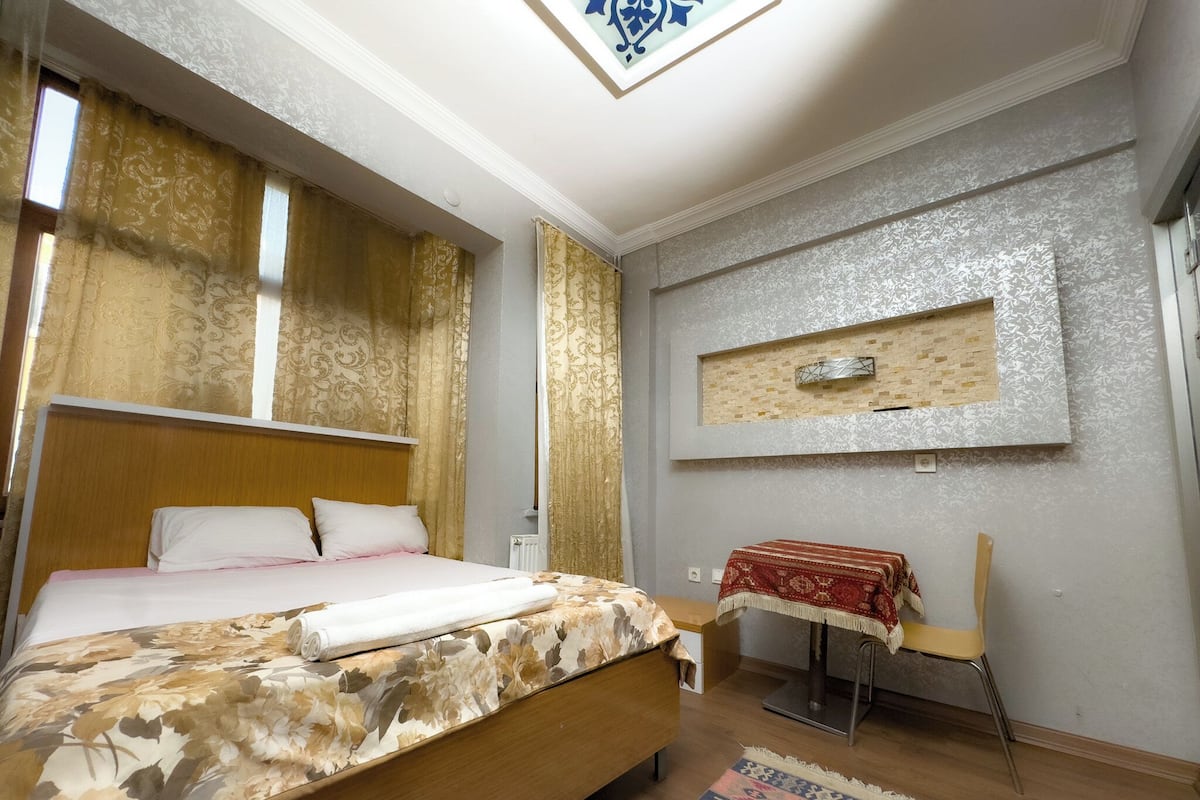 A bedroom with a bed, a chair, a small table, a patterned rug, and a decorative wall piece.