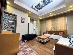 A living room with a sofa, a coffee table, and a TV. There is a kitchen visible through an open door. - The New Season Apart (Istanbul)
