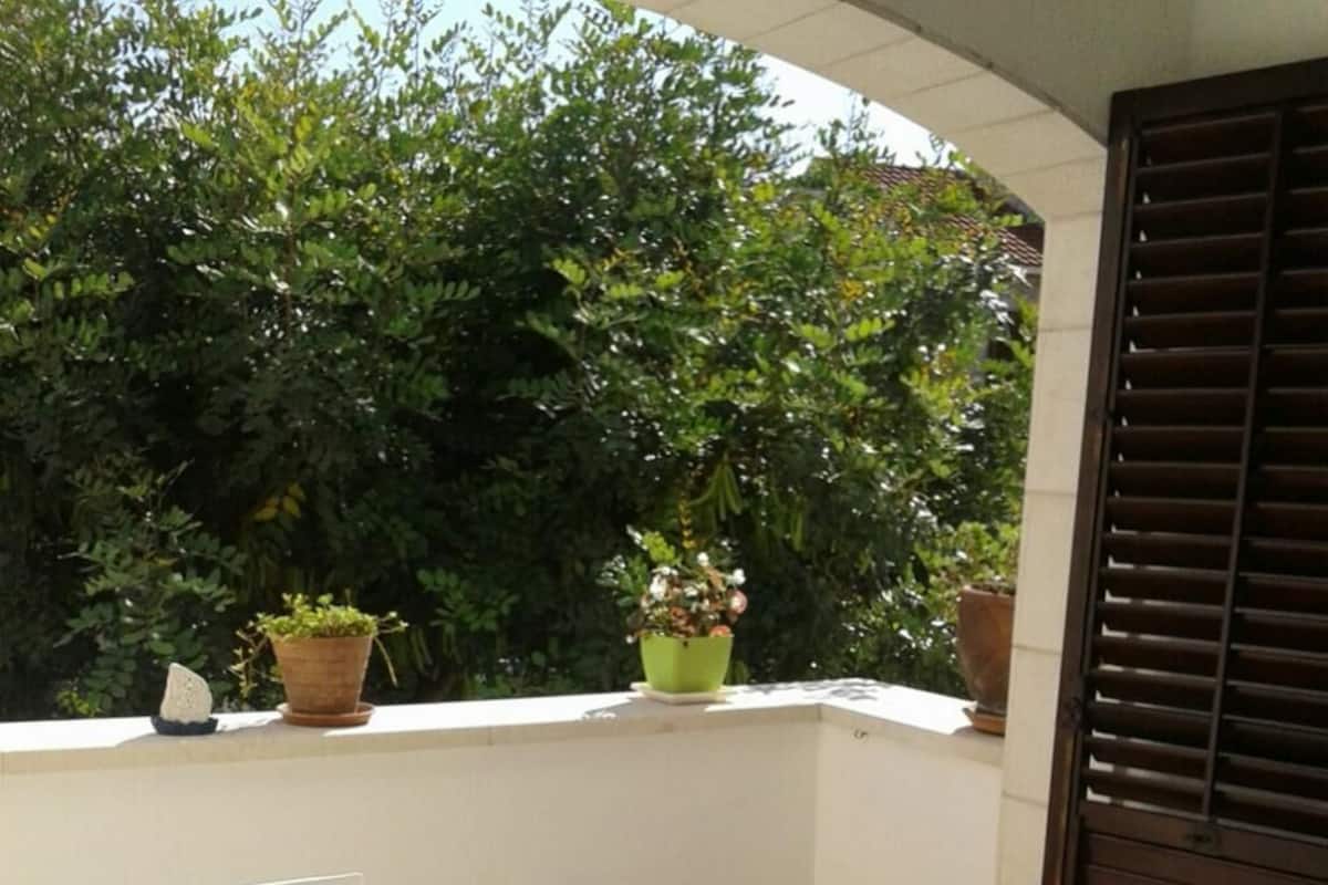 A balcony with a table covered in a blue and white tablecloth, surrounded by white chairs. There are potted plants on the balcony.