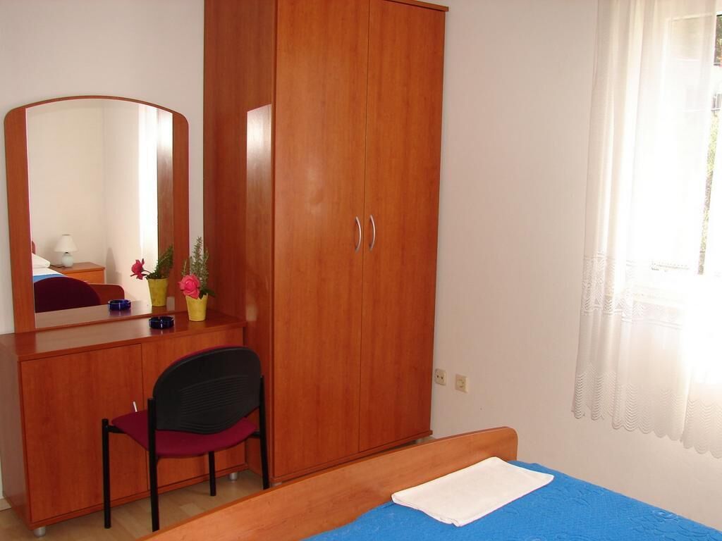 A bedroom with a wooden wardrobe, a desk with a chair, a bed with a blue bedspread, and a window with curtains.