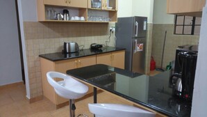 A kitchen with a black countertop, two white bar stools, a refrigerator, and tiled walls. - Scenic View Apartments Kilimani (Nairobi)