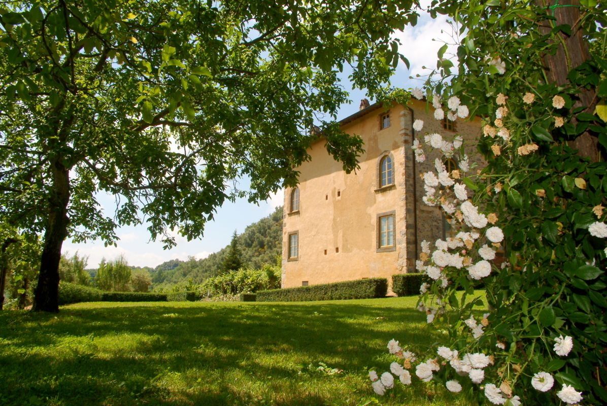 A large, two-story building with a stone facade, surrounded by lush greenery and flowering plants.