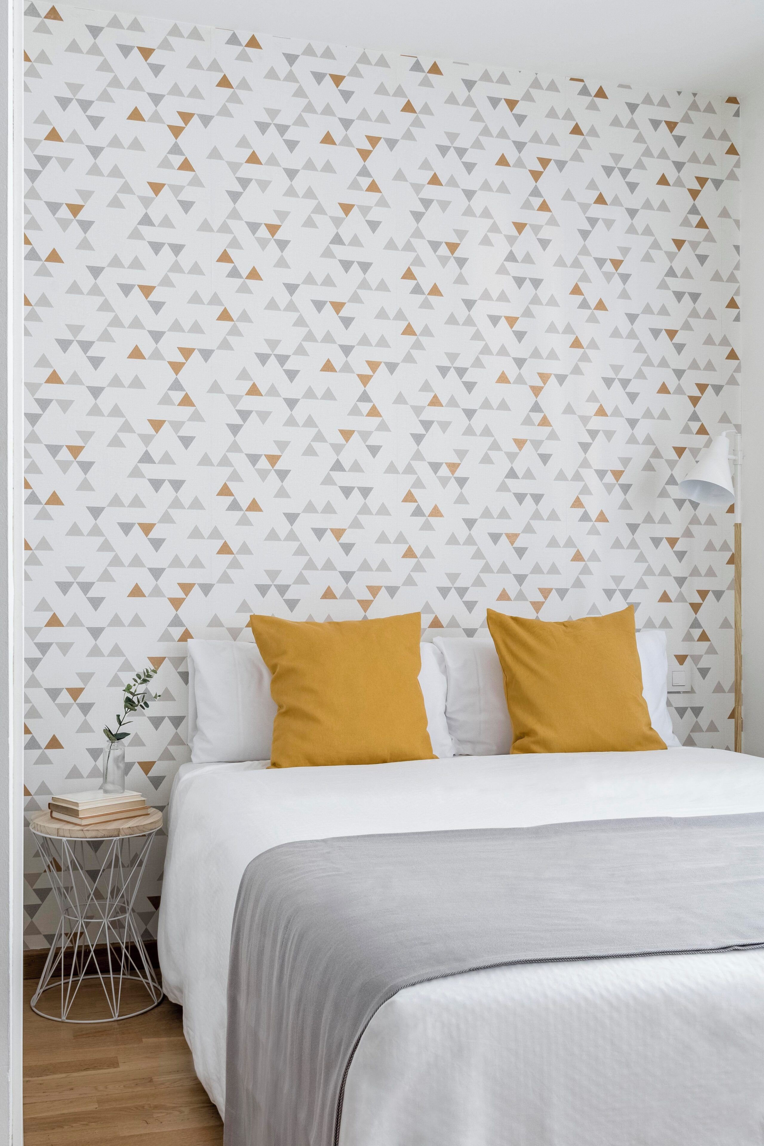 A bedroom with a patterned wallpaper, a bed with white and mustard pillows, and a small side table with a plant.