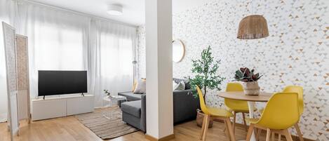 A modern living room with a flat-screen TV, grey sofa, yellow chairs, and a dining table.