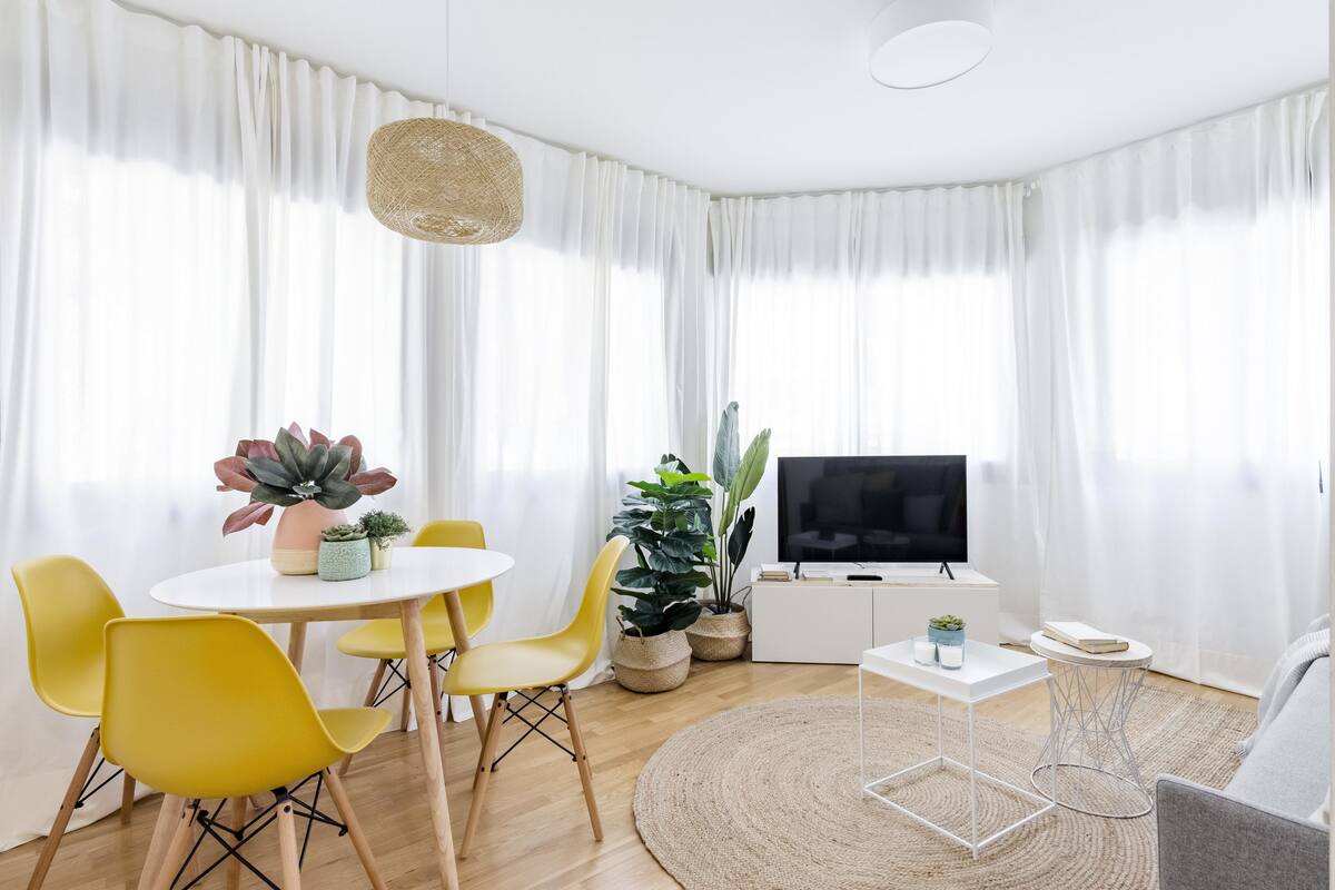A modern living room with a round dining table, yellow chairs, a TV, and potted plants.