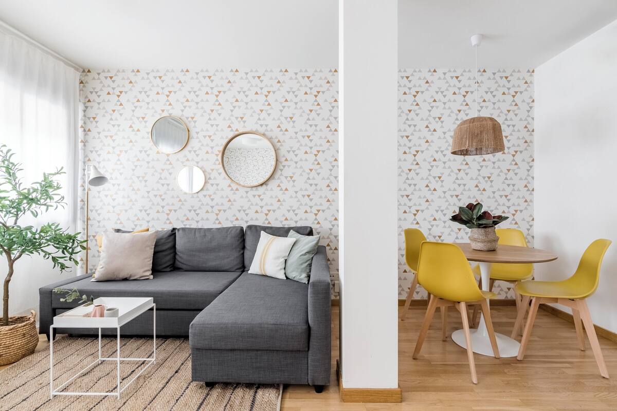 A modern living room with a grey sectional sofa, a small white table, and a dining area with yellow chairs.