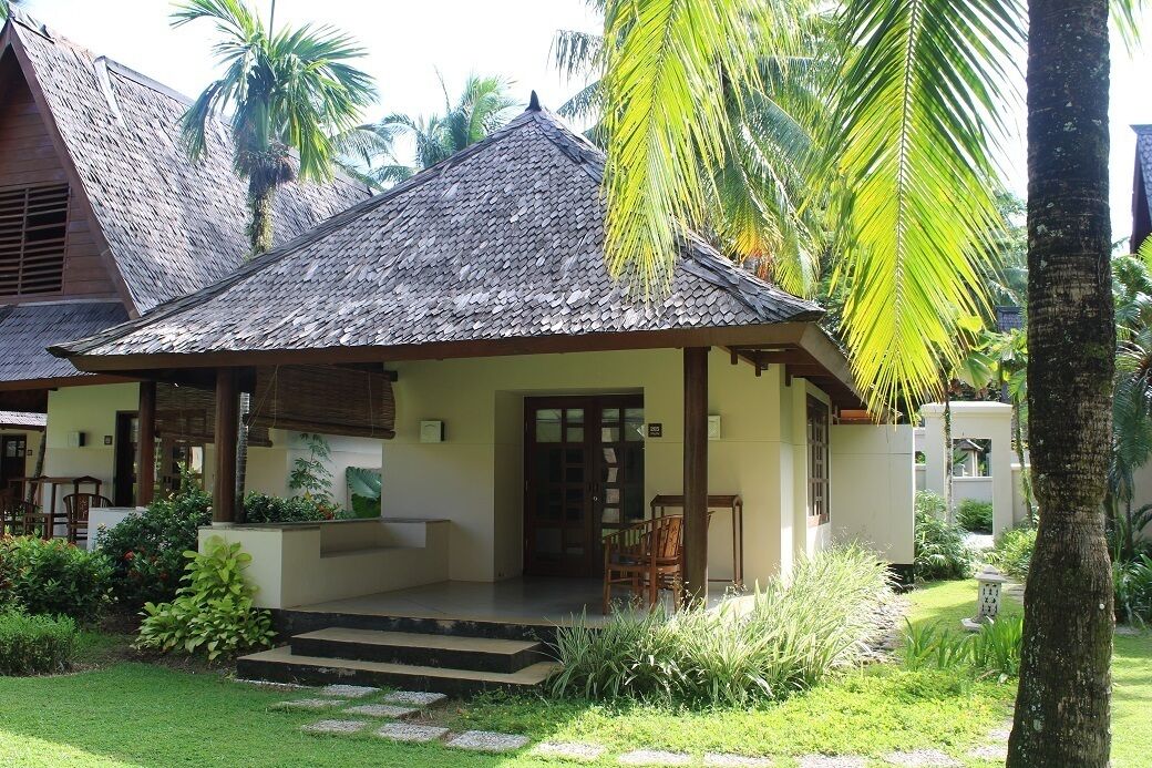 A traditional house with a thatched roof, a porch with chairs, and a well-maintained garden with palm trees and plants.