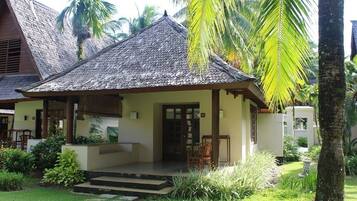 A traditional house with a thatched roof, a porch with chairs, and a well-maintained garden with palm trees and plants.