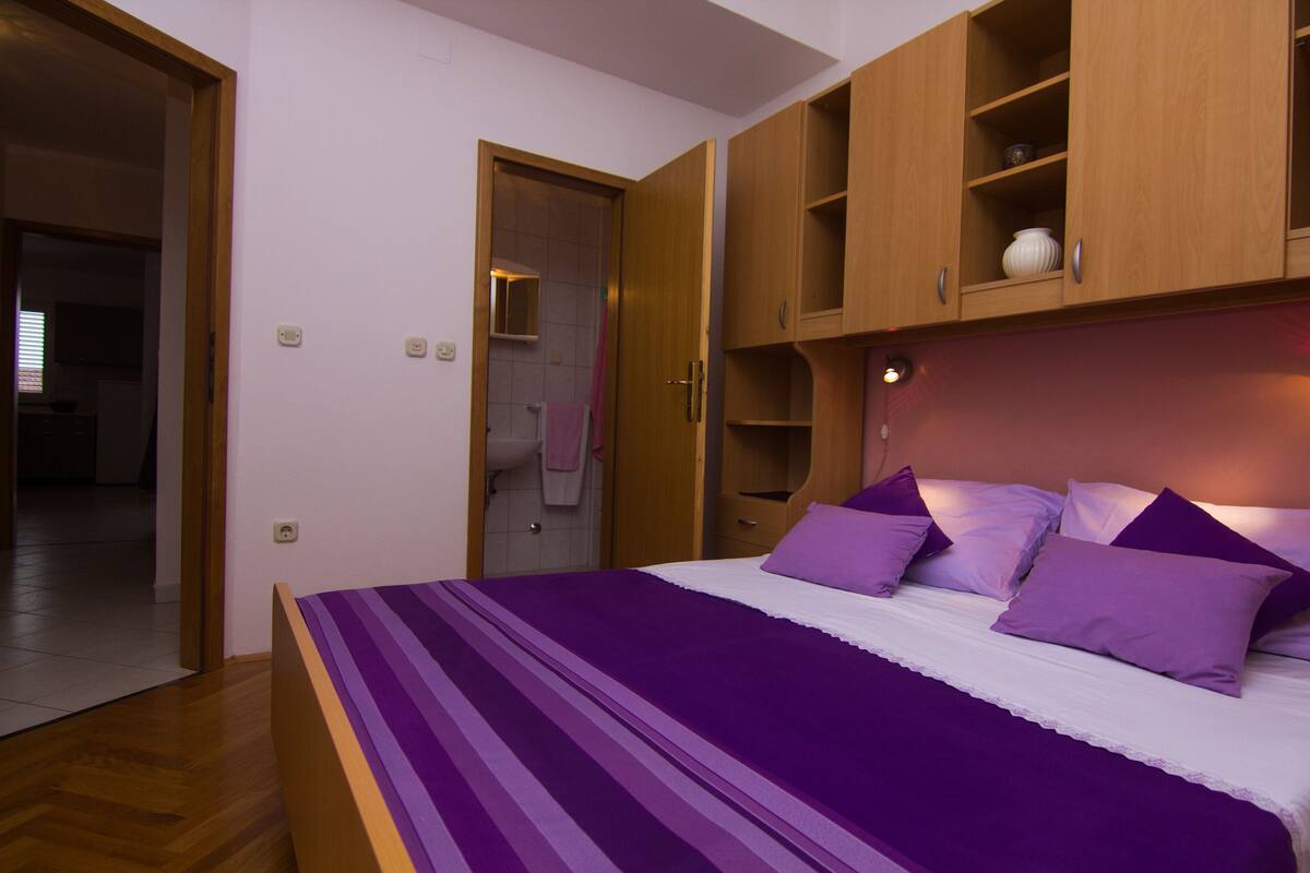 Ein Schlafzimmer mit einem Bett, einem Holzschrank und einem durchgehenden Türrahmen einsehbaren Badezimmer.
