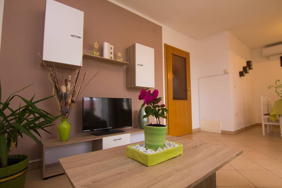 Ein modernes Wohnzimmer mit Flachbildfernseher, Couchtisch aus Holz und einer grünen Vase mit Blumen.