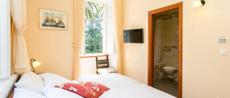 A bedroom with a bed, a window with curtains, a painting, a television, and a bathroom visible through an open door.