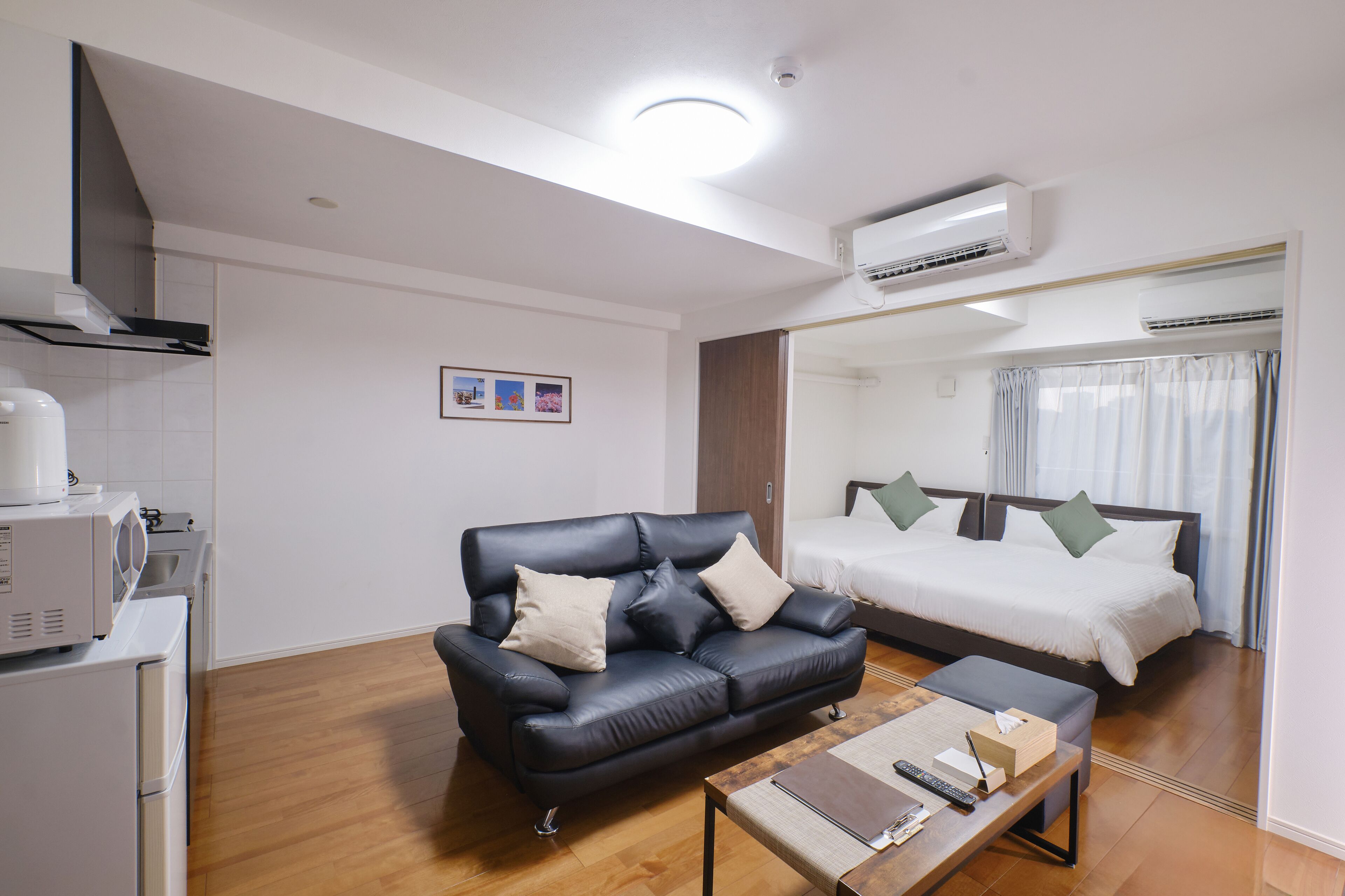 A modern hotel room with a black leather sofa, a wooden coffee table, a bed with white bedding, and a kitchenette area.