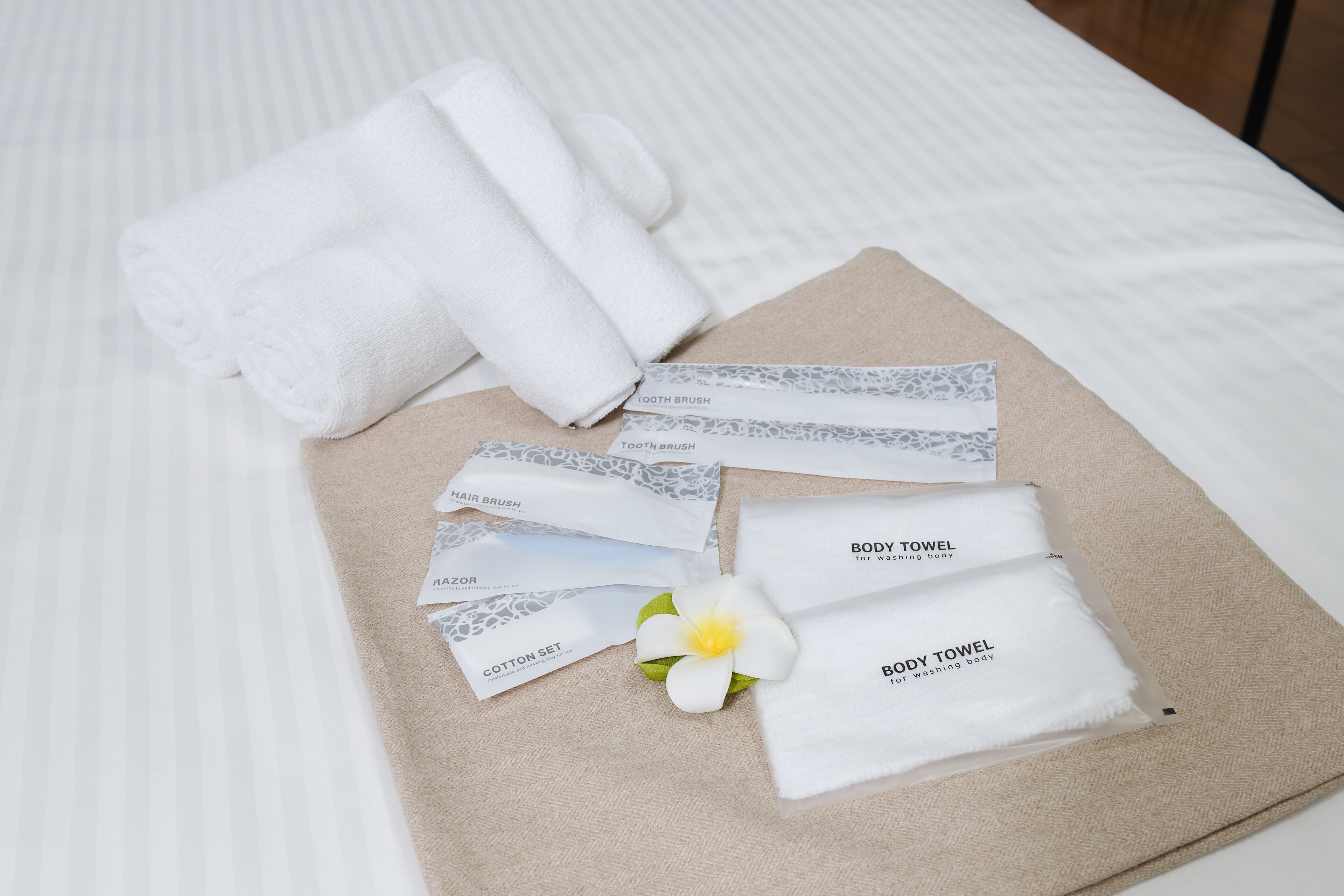 Netjes gerangschikte toiletartikelen op een beige doek, waaronder een handdoek, haarborstel, scheermes en wattorontjes, op een bed met een wit beddengoed met strepen.