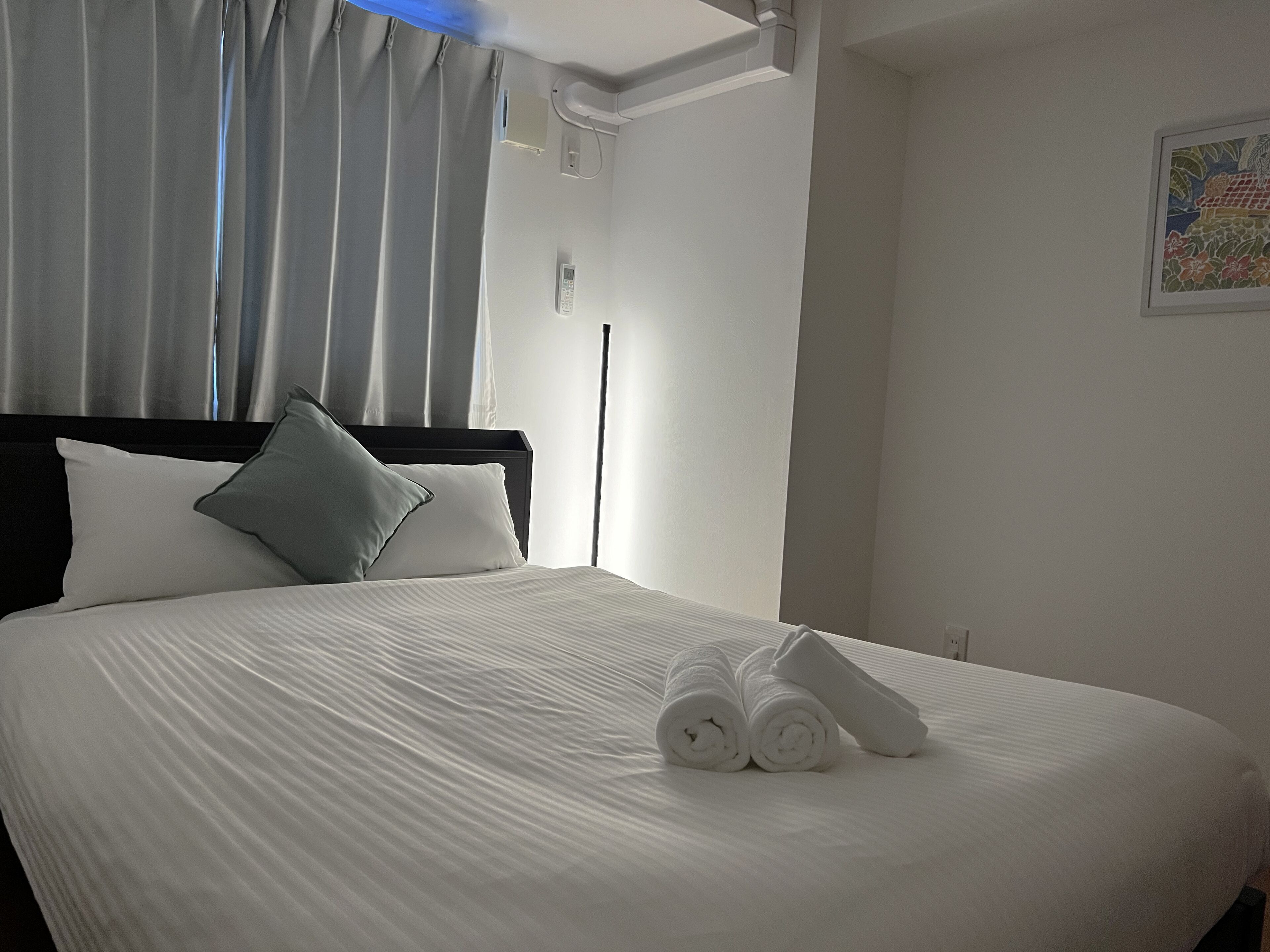 A neatly made bed with white linens, a grey pillow, and rolled towels on a white headboard.