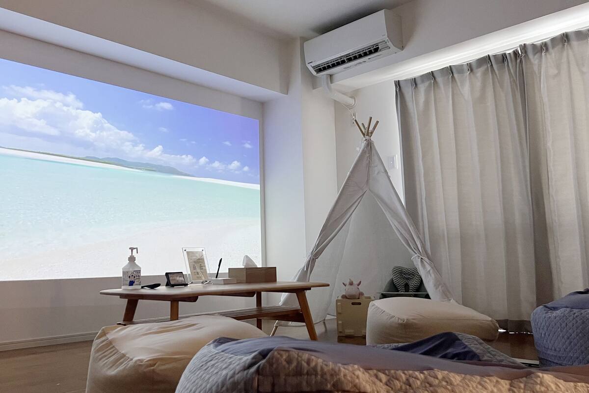 Habitación con una gran pantalla que muestra una escena de playa, una mesa de madera con detalles, una estructura tipo tienda y una cama con vistas.