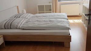 A single bed with a wooden frame and a white mattress, a bedside table, and a white cabinet in a room with wooden flooring and a radiator. - Zur schönen Aussicht (Hellenthal)