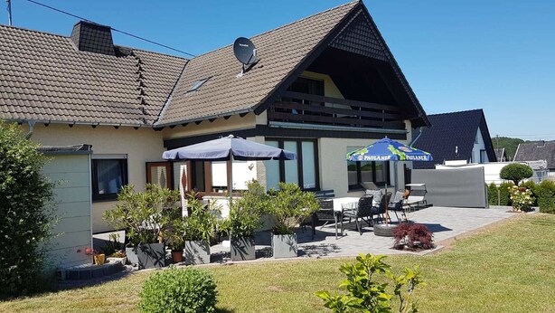 A two-story house with a sloped roof, a satellite dish, outdoor seating with umbrellas, and potted plants. - Zur schönen Aussicht (Hellenthal)