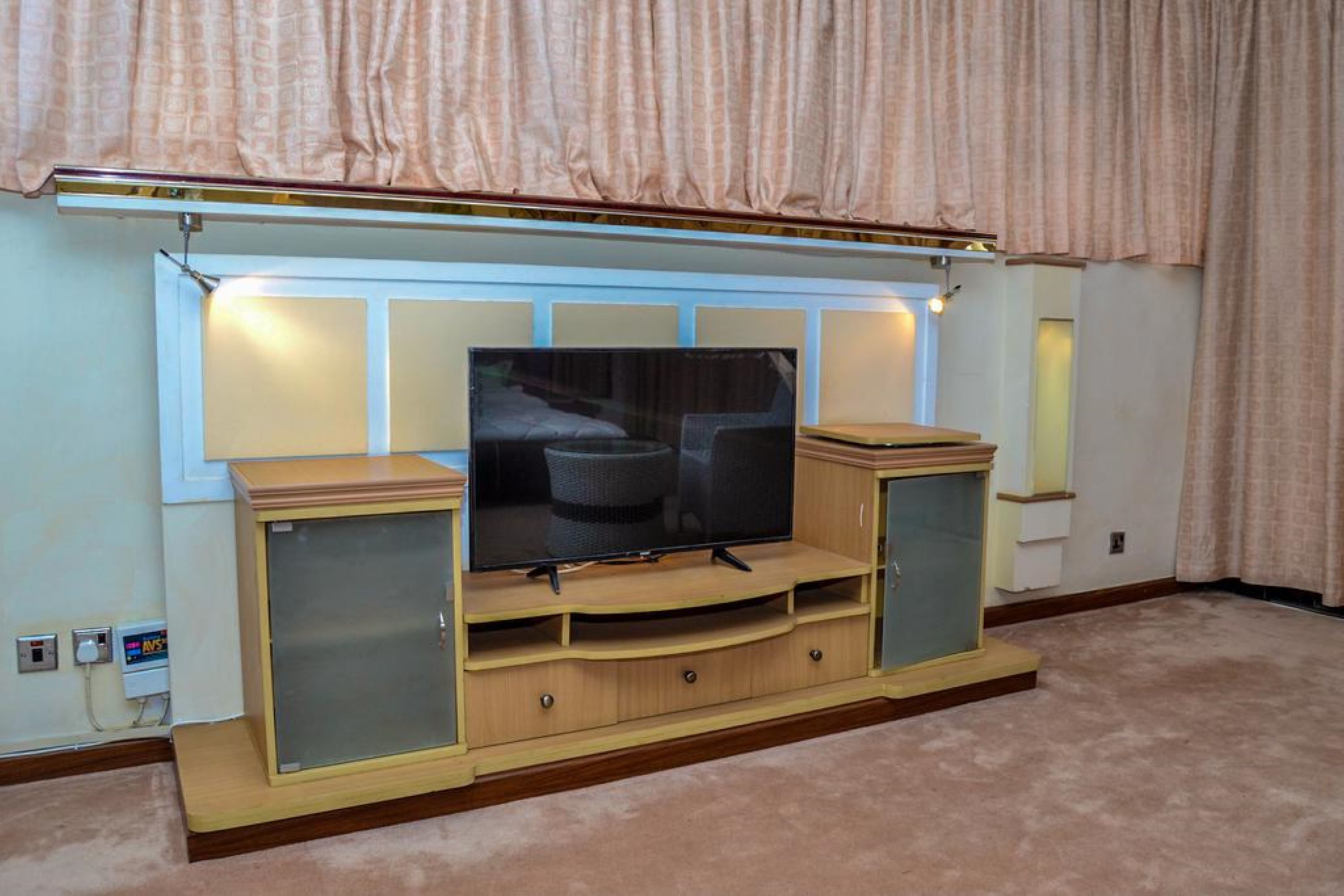 A television set on a wooden entertainment center with a glass front, located in a room with beige carpet and light-colored walls.