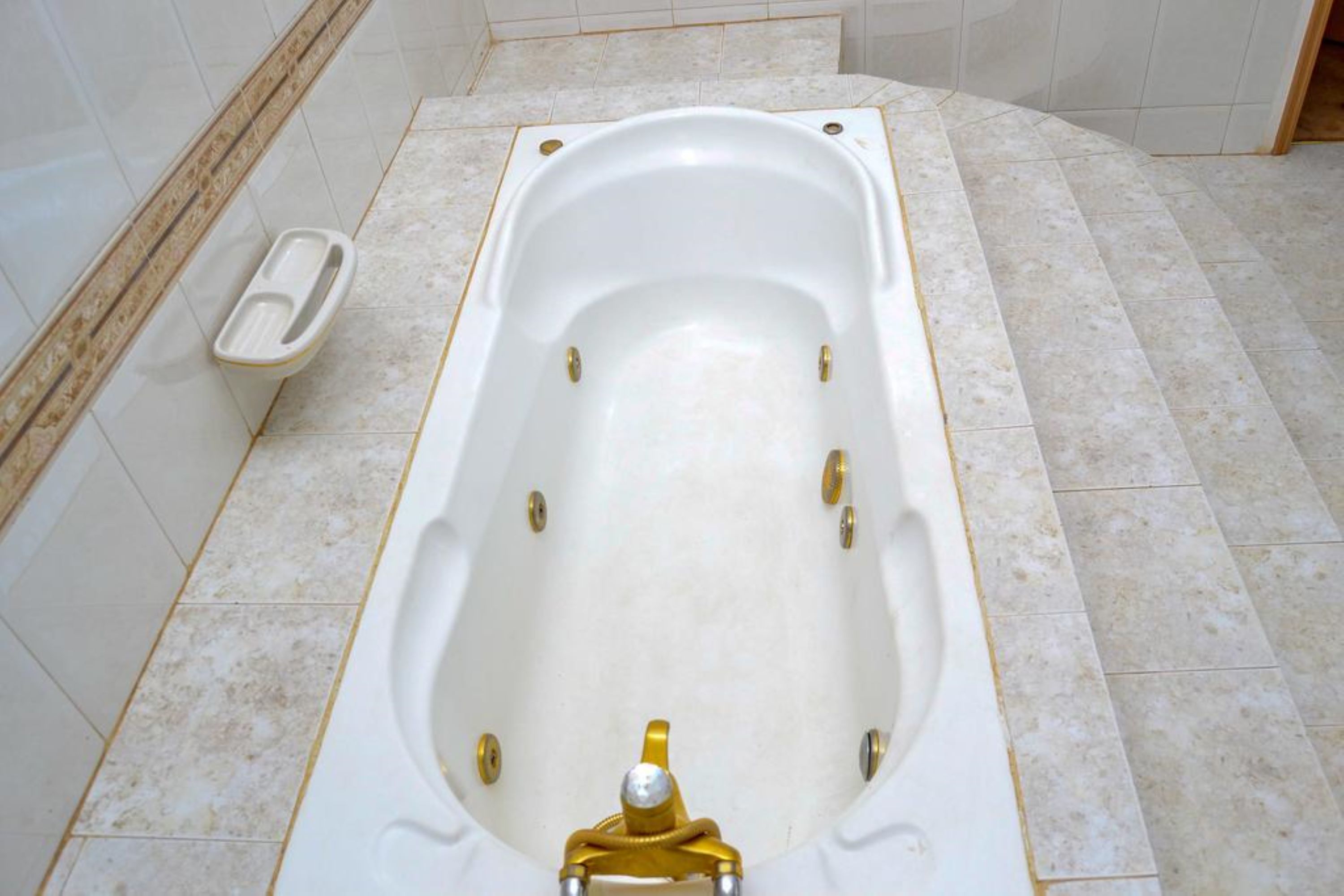 Una bañera grande y rectangular con un grifo dorado en un baño con paredes y suelo de baldosas.