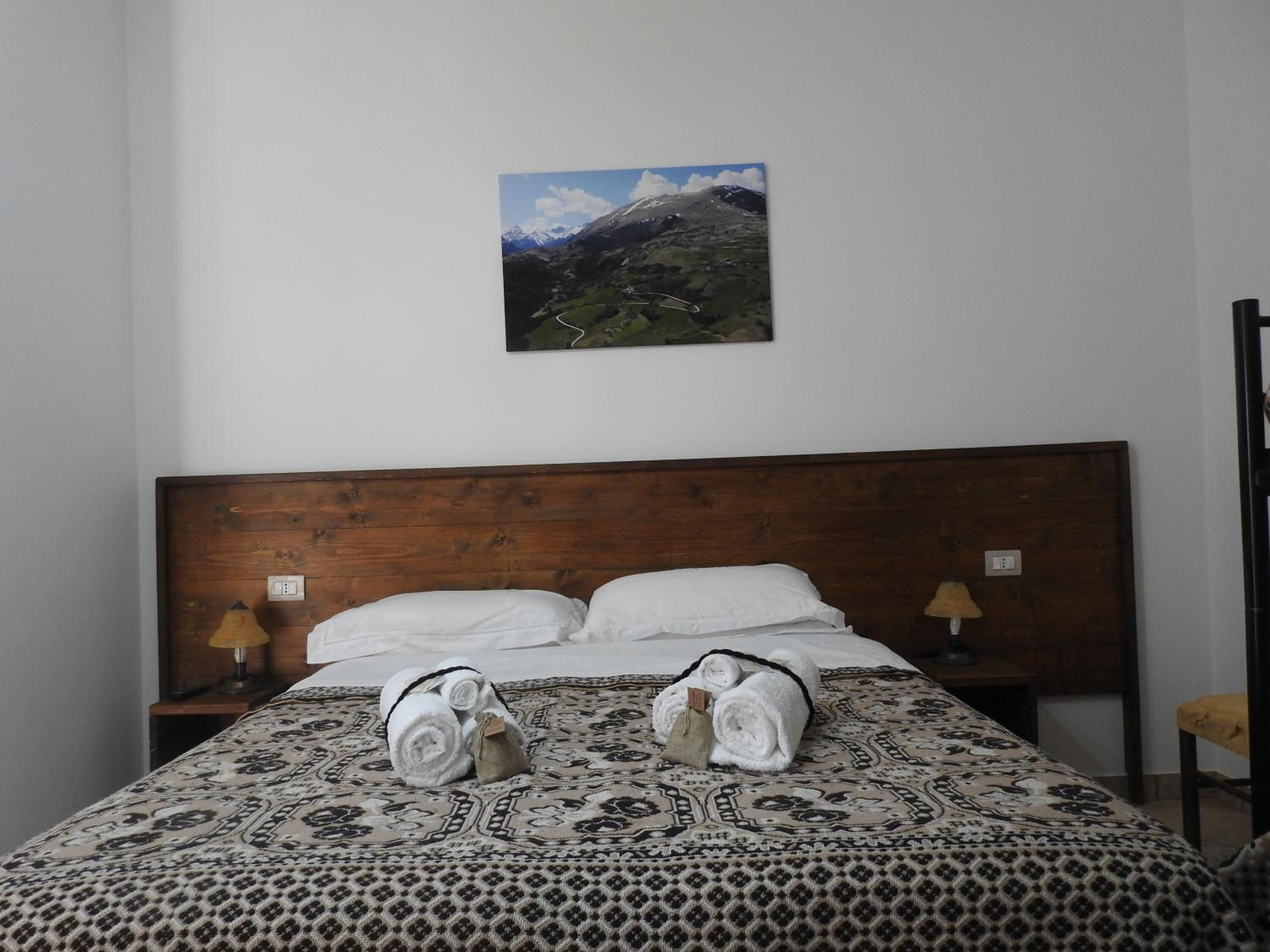 Quarto de hotel com cabeceira de madeira, duas mesas de cabeceira com luminárias, colcha estampada e pintura de paisagem montanhosa na parede.