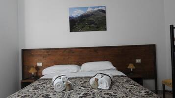 A hotel room with a wooden headboard, two bedside tables with lamps, a patterned bedspread, and a mountain landscape painting on the wall.