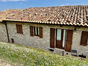 Un edificio de piedra con techo de tejas, contraventanas de madera y un pequeño jardín con flores blancas y amarillas.