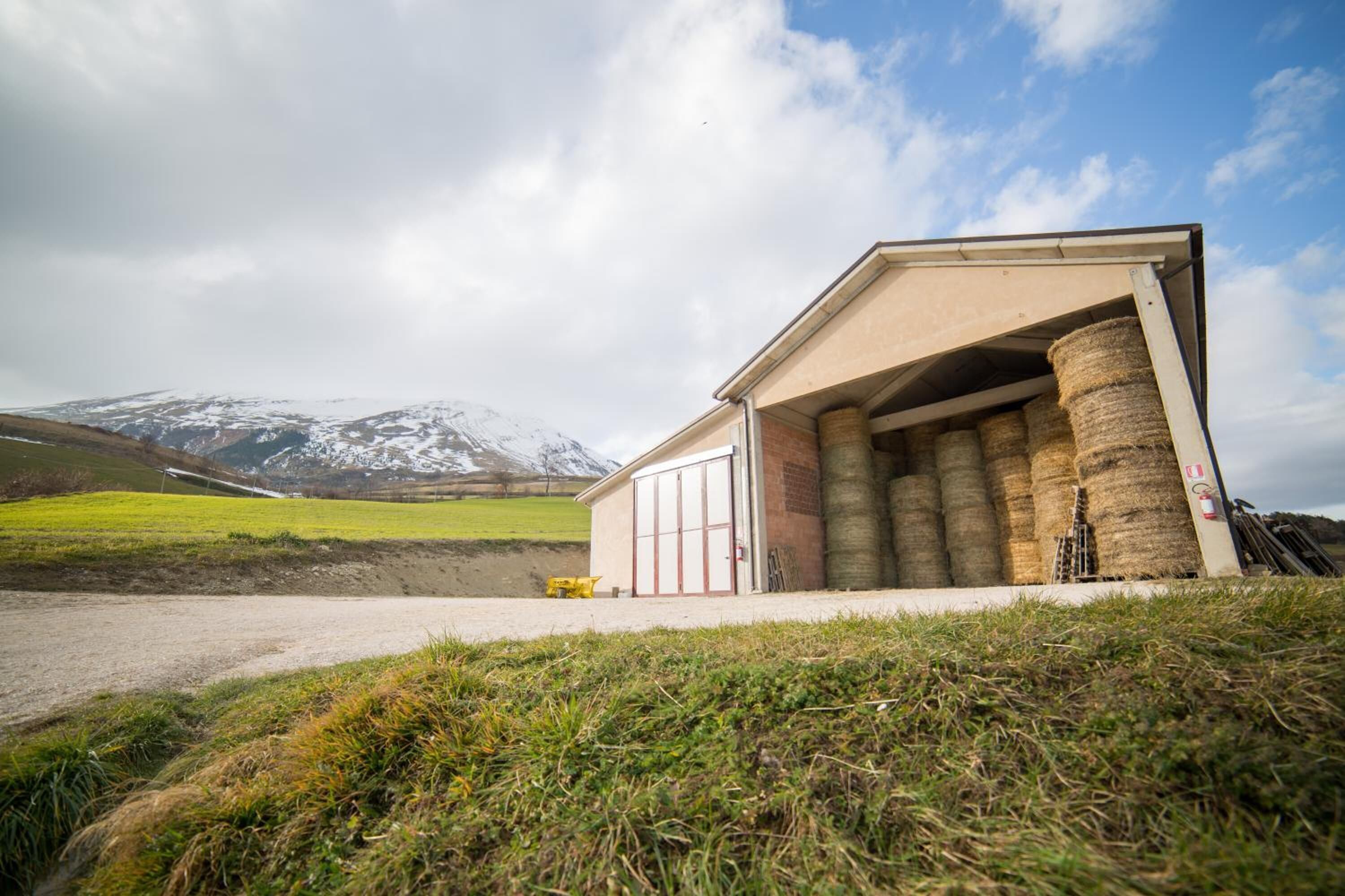 Un bâtiment doté d’une grande porte de garage ouverte, des balles de foin entreposées à l’intérieur, et un paysage montagneux en arrière-plan.