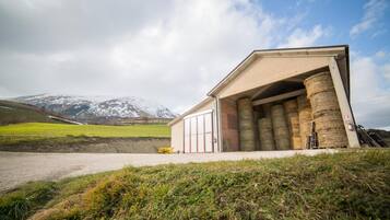Un bâtiment doté d’une grande porte de garage ouverte, des balles de foin entreposées à l’intérieur, et un paysage montagneux en arrière-plan.