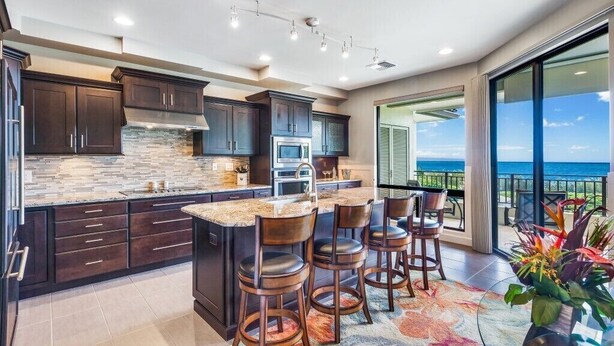A modern kitchen with dark wood cabinets, a large island with bar stools, and a view of the ocean through large windows. - ONE of Our BEST! Fresh Pricing, UPSCALE Renovations; (22V-1) where you get HUGE OCEAN Views (Lahaina)