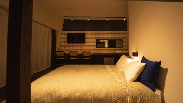 Ein Schlafzimmer mit einem Bett, einem Schreibtisch und einem Stuhl. Ein Fenster mit Vorhängen und eine Ziegelwand befinden sich hinter dem Schreibtisch.