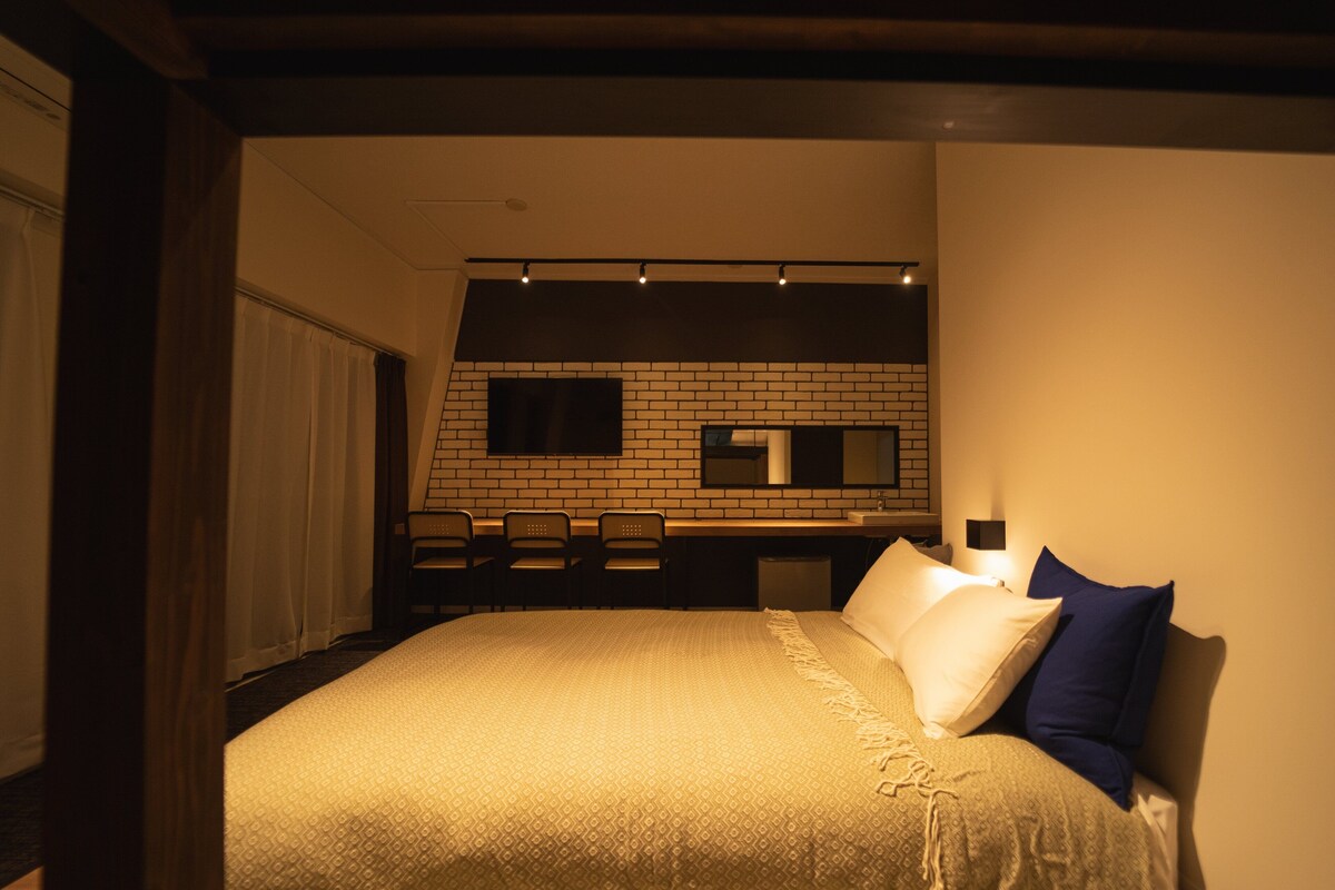 Une chambre à coucher avec un lit, un bureau et une chaise. Il y a une fenêtre avec des rideaux et un mur en briques derrière le bureau.