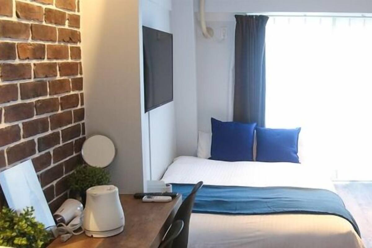 Een compacte hotelkamer met een bakstenen muur, een bed met blauwe kussens, een stapelbed, een televisie en een klein tafeltje met een waterkoker.