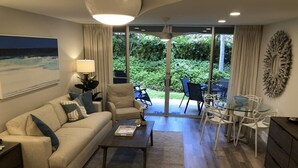 Ein modernes Wohnzimmer mit Sofa, Couchtisch und Essbereich. Ein großes Fenster bietet Blick auf Grünflächen.