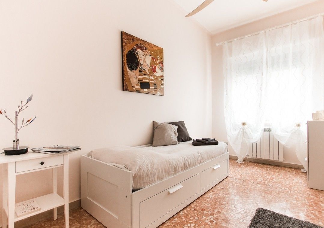 A minimalist bedroom with a bed, a bedside table, a radiator, and a window with sheer curtains.