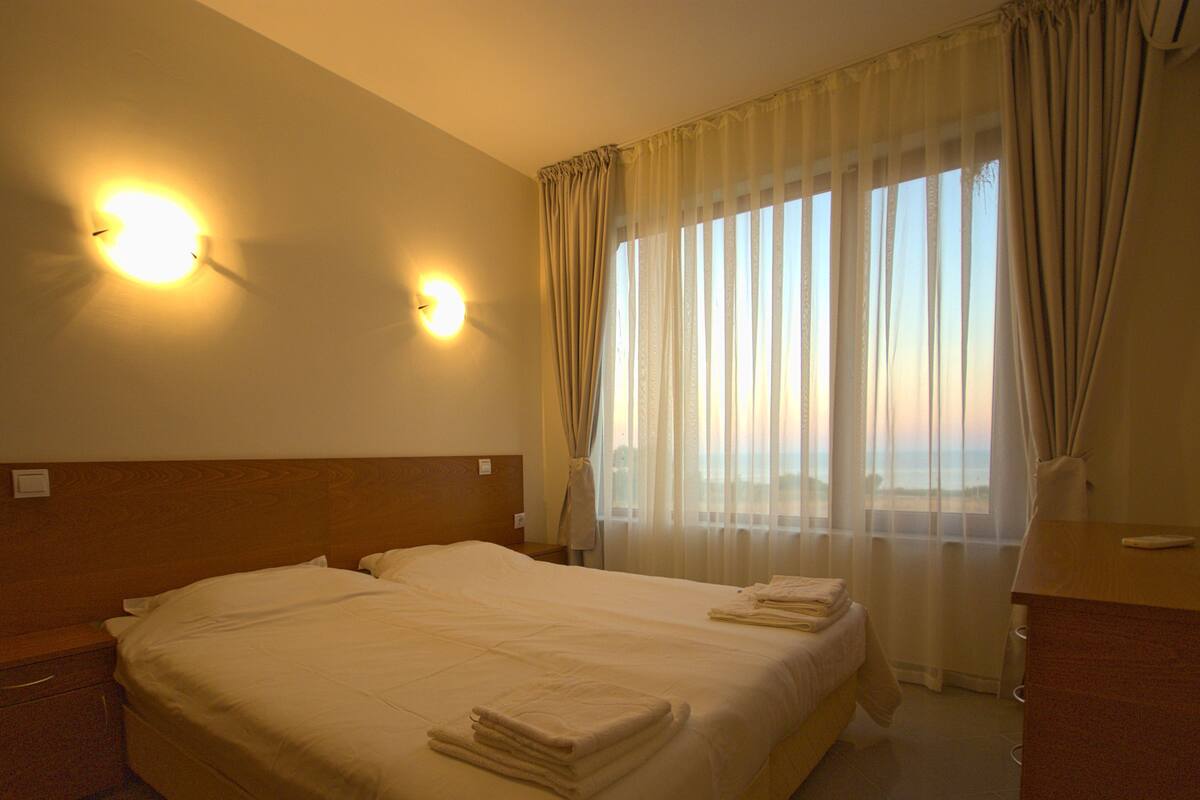 A hotel room with a large bed, two wall-mounted lights, a window with curtains, and a bedside table with a lamp.
