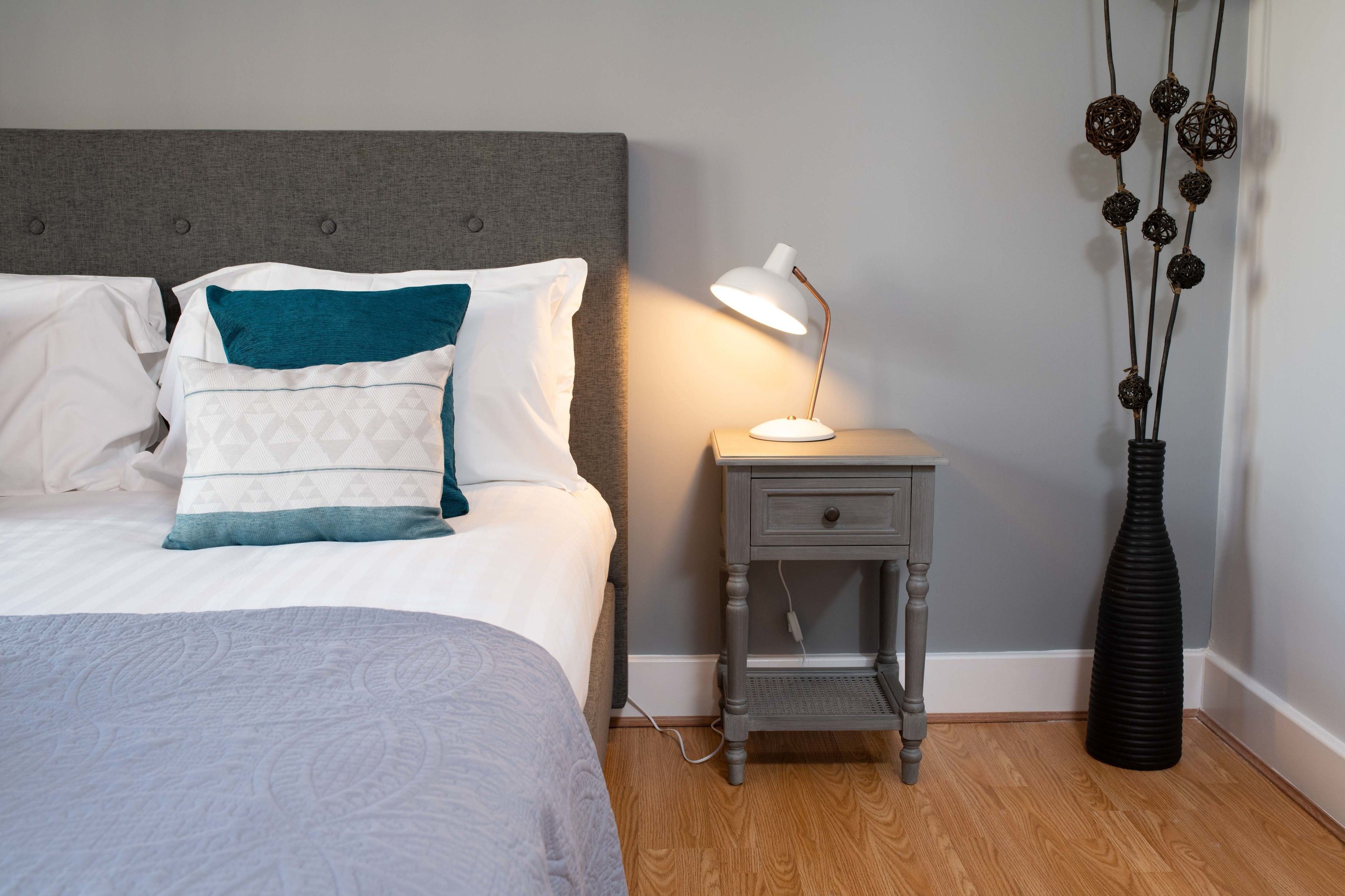A neatly made bed with patterned bedding, a bedside table with a lamp, and a decorative vase with hanging ornaments.